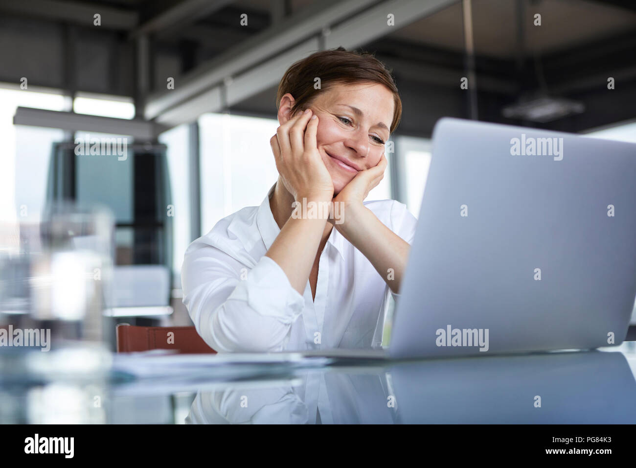 Smiling businesswoman sitting at glass table in office looking at laptop Stock Photo