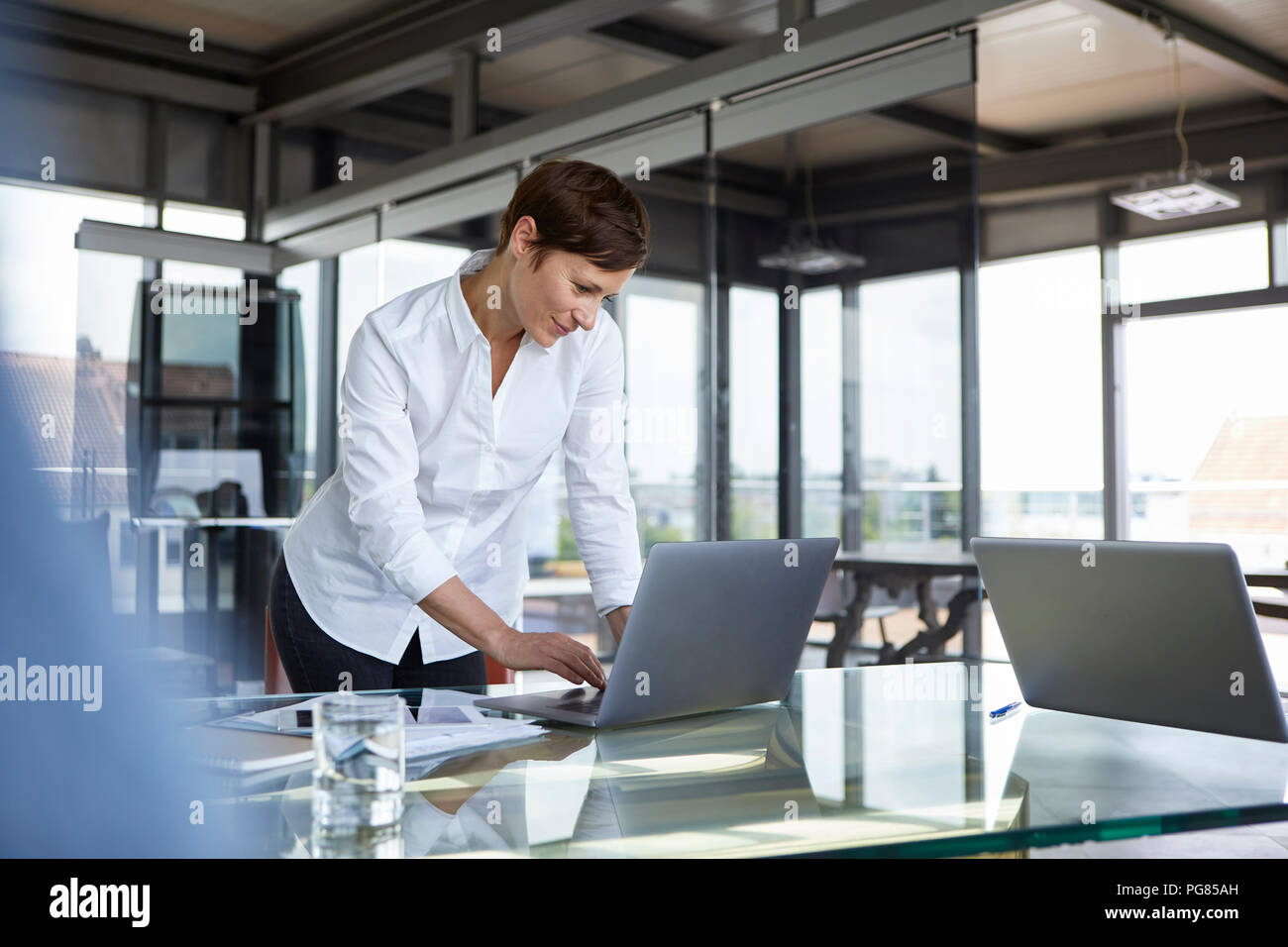 Businesswoman standing at glass table in office using laptop Stock Photo
