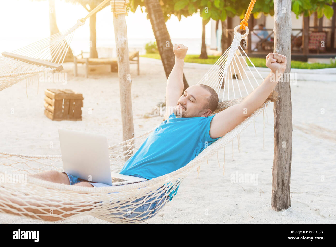 The man quite stretches lying in a hammock with a laptop. The concept of remote work. Stock Photo
