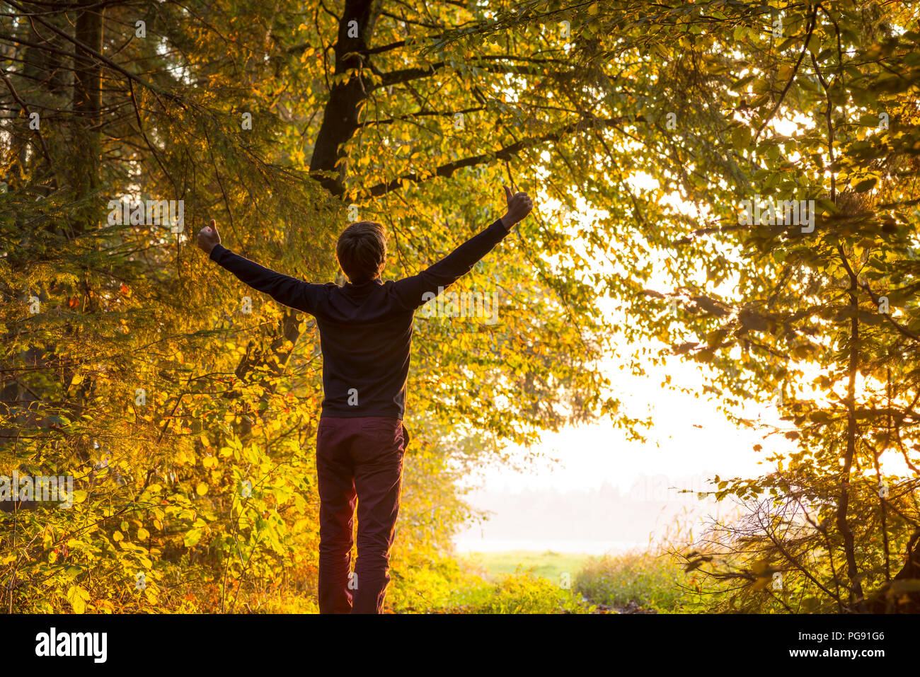 Young man standing on the edge of forested area raising his arms in celebration turned towards a bright sunlight waiting for him just outside the fore Stock Photo