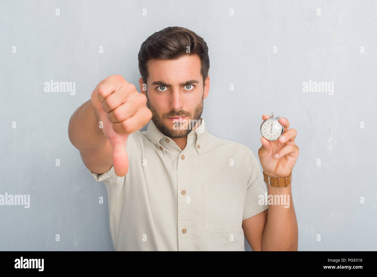 Handsome young man over grey grunge wall holding stopwatch with angry ...