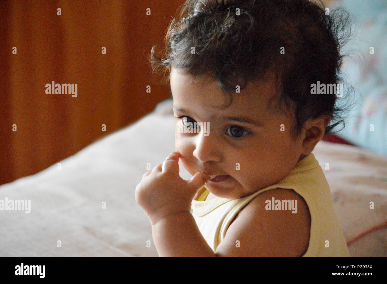 Infant in yellow top sitting on bed in a deep thoughtful expression with her little thumb around her nose and index finger touching her right cheek Stock Photo