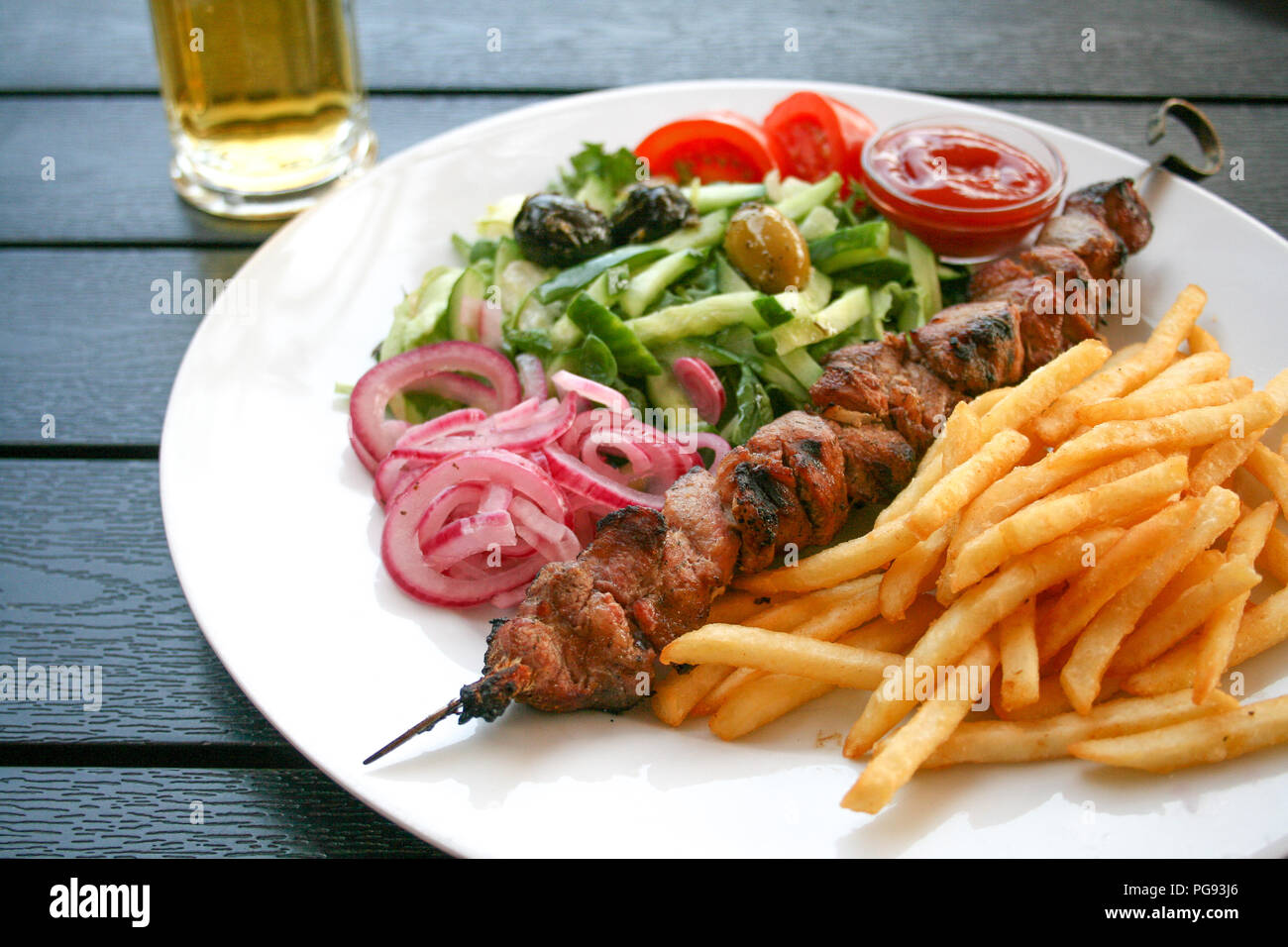 Shashlik or shashlyk of pork, dish of skewered and grilled cubes of meat, with red onion, vegetables, tomatoes, olives and french fries on a wooden ta Stock Photo