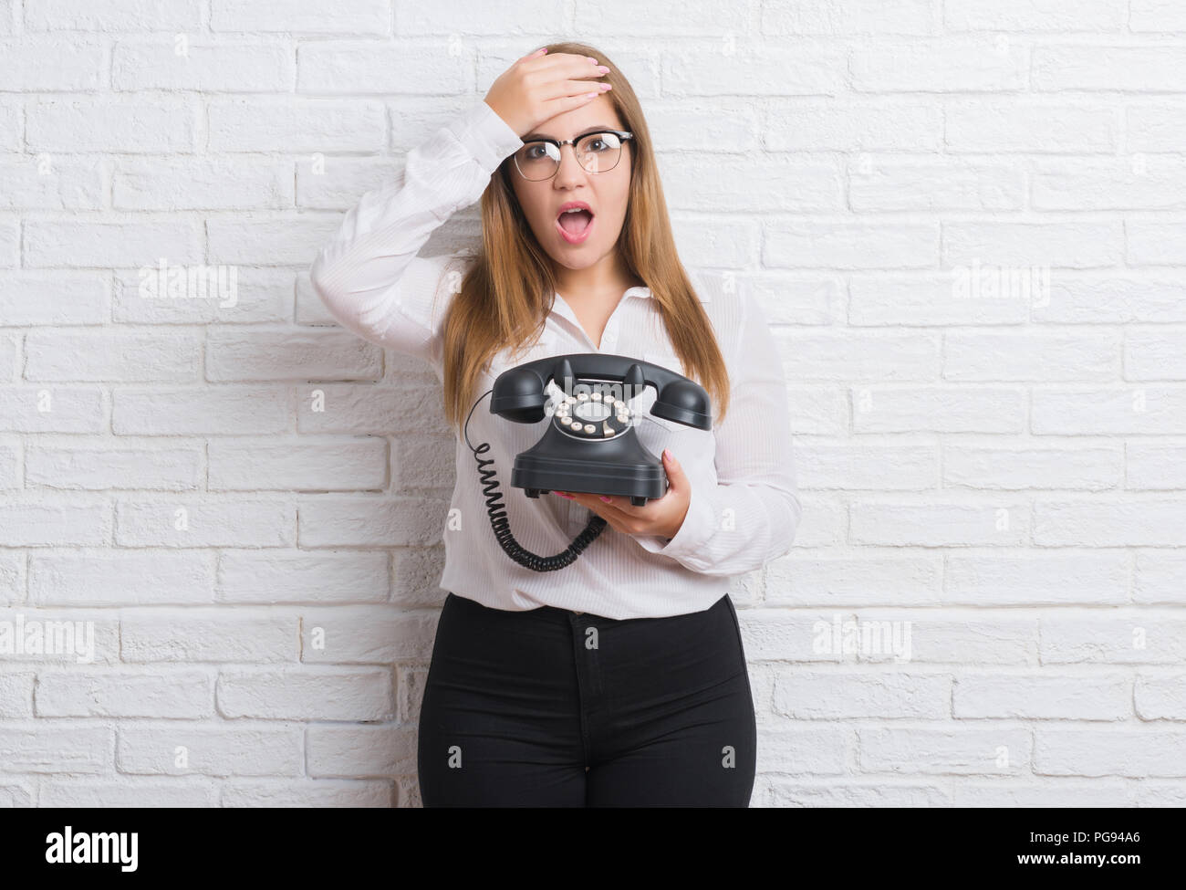 Young adult woman over white brick wall holding vintage telephone stressed with hand on head, shocked with shame and surprise face, angry and frustrat Stock Photo