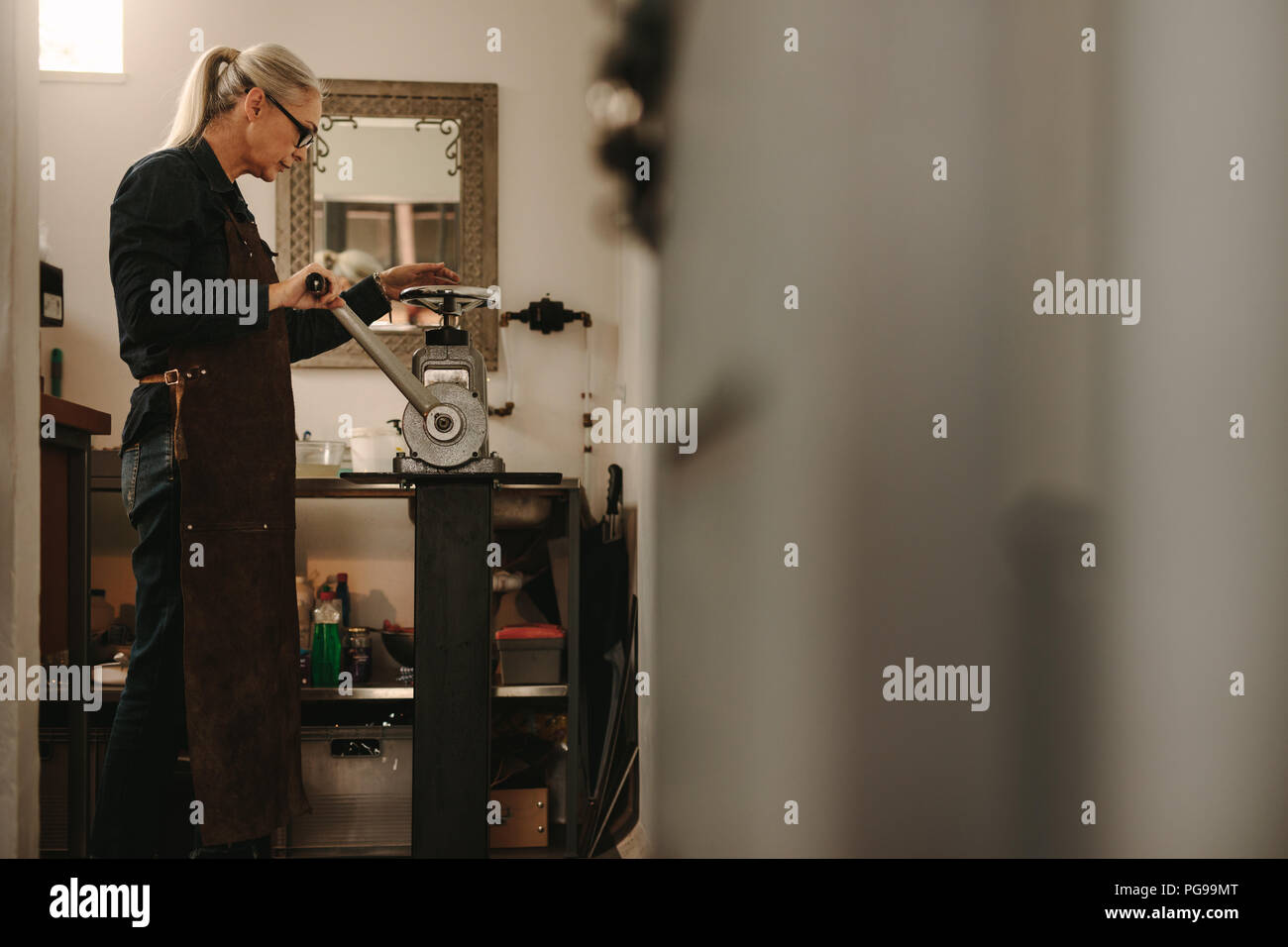 Senior female goldsmith crafting metal on rolling machine in workshop. Mature woman working in goldsmith workshop. Stock Photo