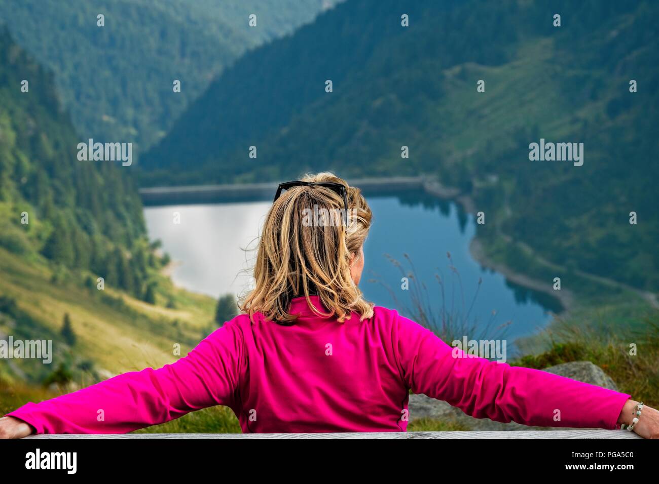 A blonde woman sitting on a bench and looking at the horizon. A mountain valley with a lake is visible in the background. Shot from behind Stock Photo