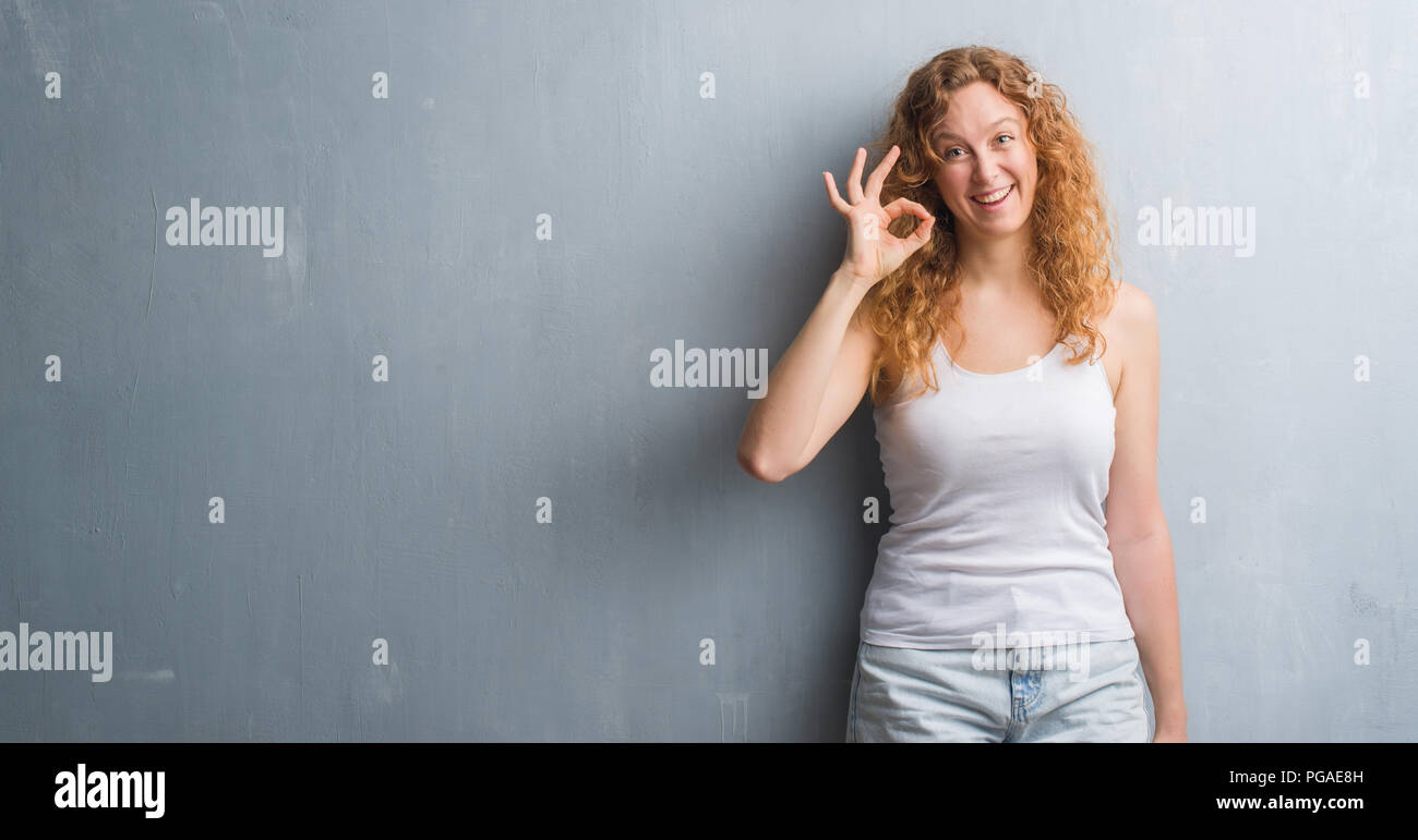 Young redhead woman over grey grunge wall doing ok sign with fingers ...