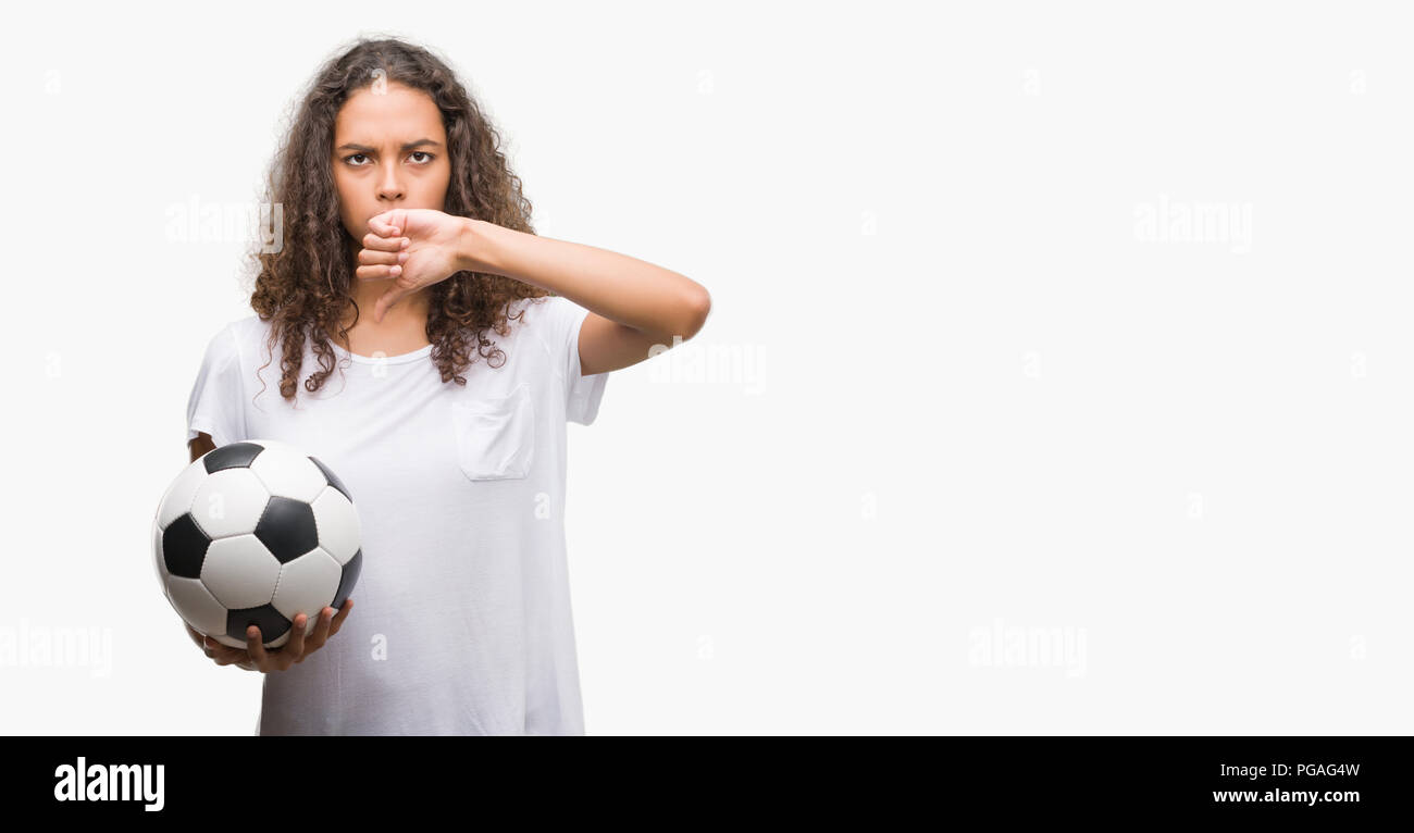 Young hispanic woman holding soccer football ball with angry face, negative sign showing dislike with thumbs down, rejection concept Stock Photo