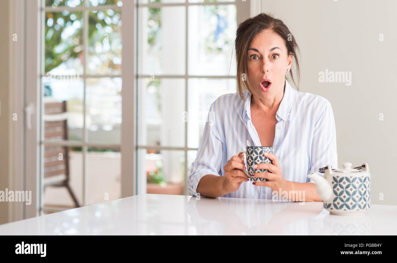 Middle aged woman drinking a cup of tea or coffee scared in shock with