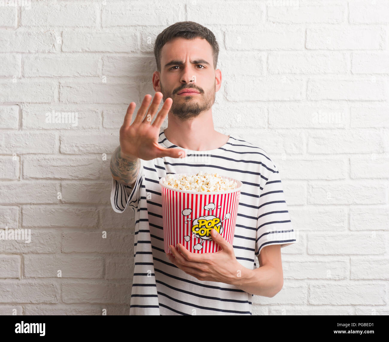 Young adult man eating popcorn standing over white brick wall with open hand doing stop sign
