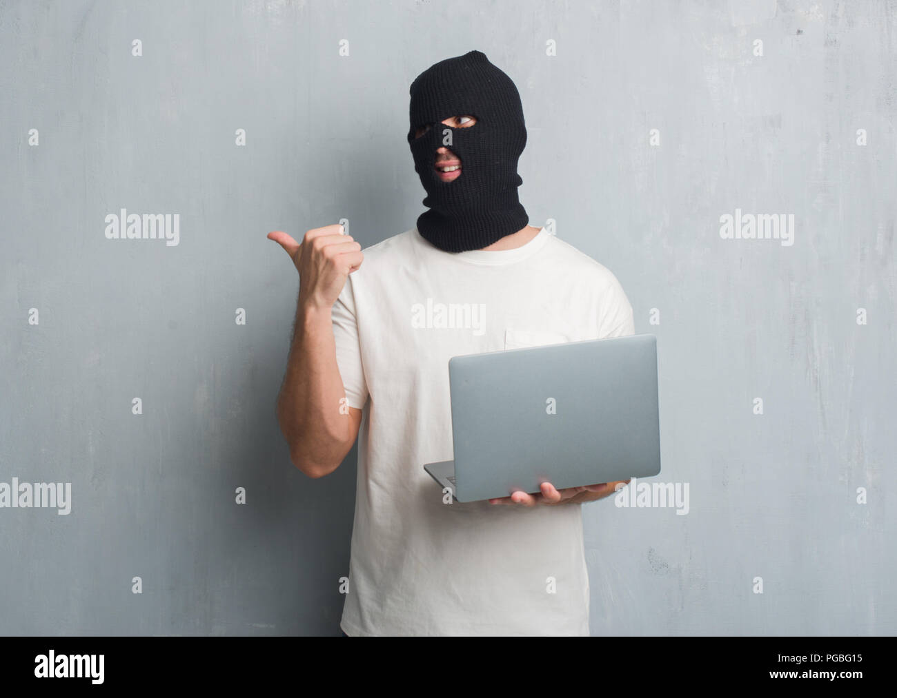 Young caucasian hacker man over grey grunge wall doing cyber attack using laptop pointing and showing with thumb up to the side with happy face smilin Stock Photo