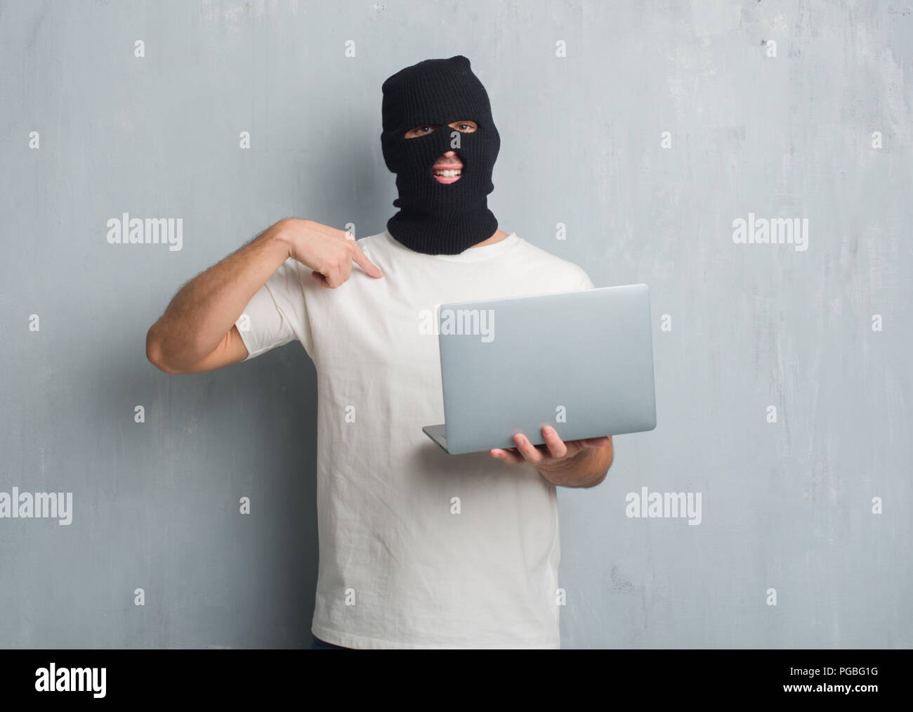 Young caucasian hacker man over grey grunge wall doing cyber attack using laptop with surprise face pointing finger to himself Stock Photo