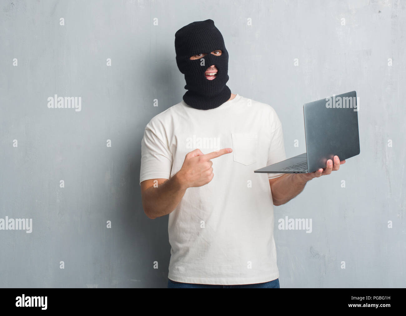 Young caucasian hacker man over grey grunge wall doing cyber attack using laptop very happy pointing with hand and finger Stock Photo