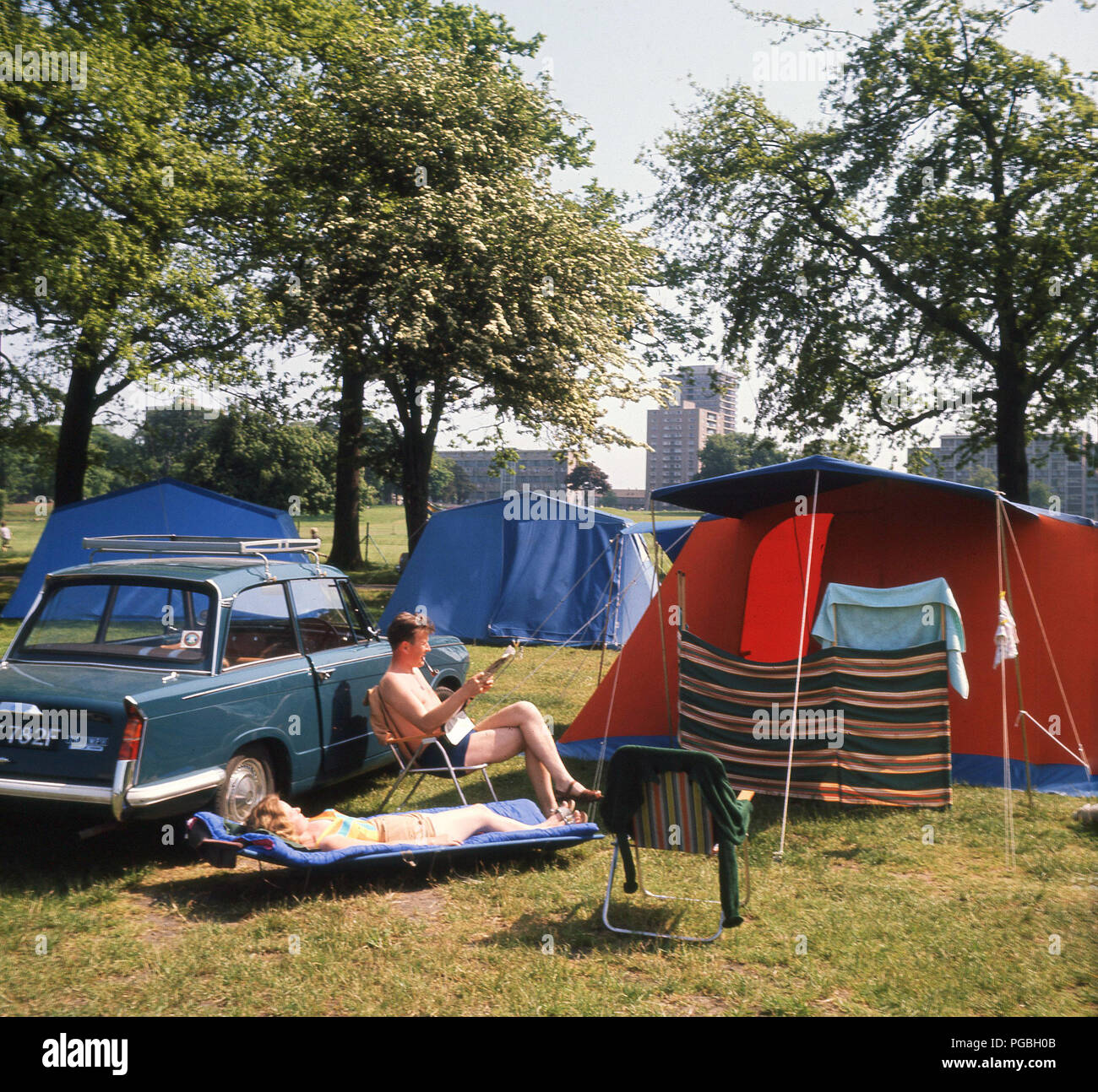1970s, A couple sitting by their car, a Triumph Herald relaxing in the sunshine by their tent on a simple camping holiday on the outskirts of the city of Edinburgh, Scotland. The man is sitting reading a newspaper, while the woman lies on a sun lounger, a low reclining chair, popular in the era for use when at the beach, in the garden or camping. Stock Photo