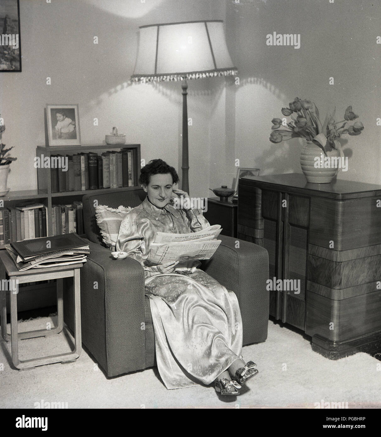 1950s, historical, picture shows a lady sitting in armchair in a lving room wearing a long dressing gown reading a newspaper, England, UK. Stock Photo