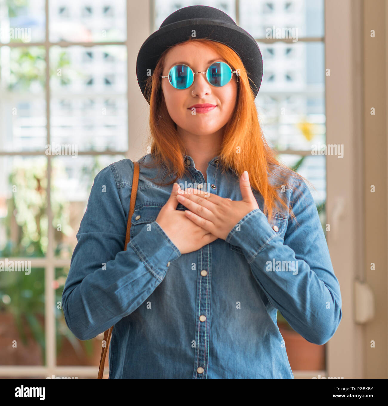 Stylish redhead woman wearing bowler hat and sunglasses smiling with hands on chest with closed eyes and grateful gesture on face. Health concept. Stock Photo