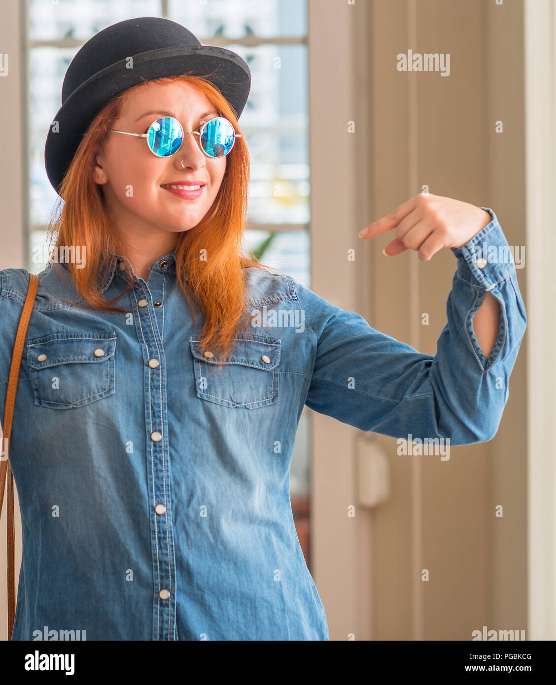 Stylish redhead woman wearing bowler hat and sunglasses looking confident with smile on face, pointing oneself with fingers proud and happy. Stock Photo