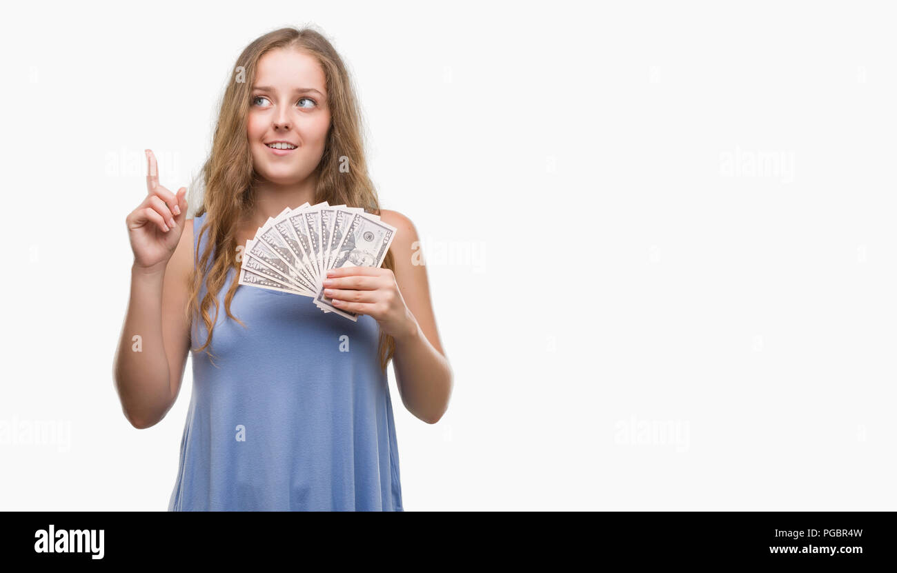 Young blonde woman holding dollars surprised with an idea or question pointing finger with happy face, number one Stock Photo
