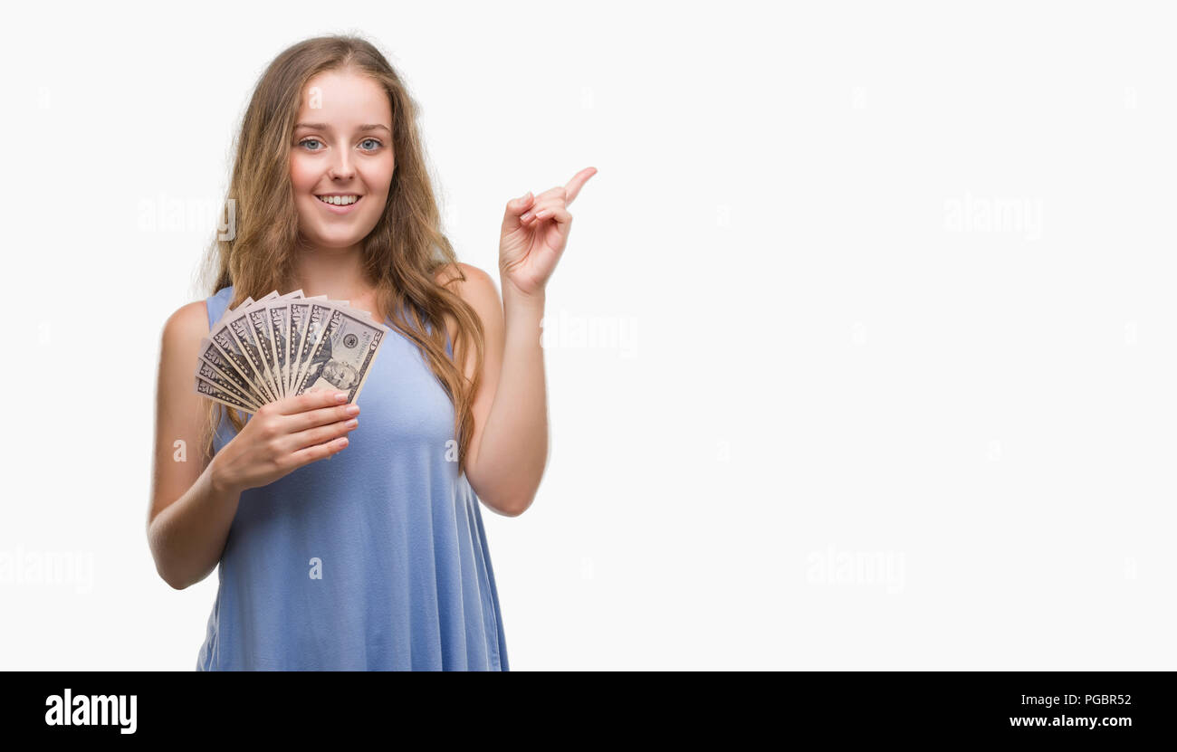 Young blonde woman holding dollars very happy pointing with hand and finger to the side Stock Photo