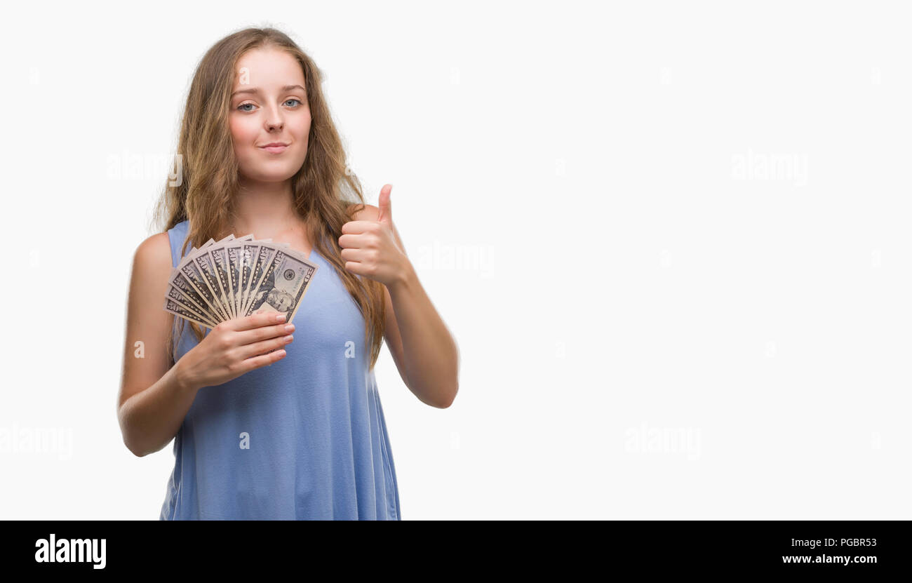 Young blonde woman holding dollars happy with big smile doing ok sign, thumb up with fingers, excellent sign Stock Photo
