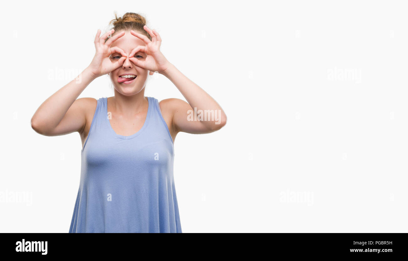 Young blonde woman doing ok gesture like binoculars sticking tongue out, eyes looking through fingers. Crazy expression. Stock Photo