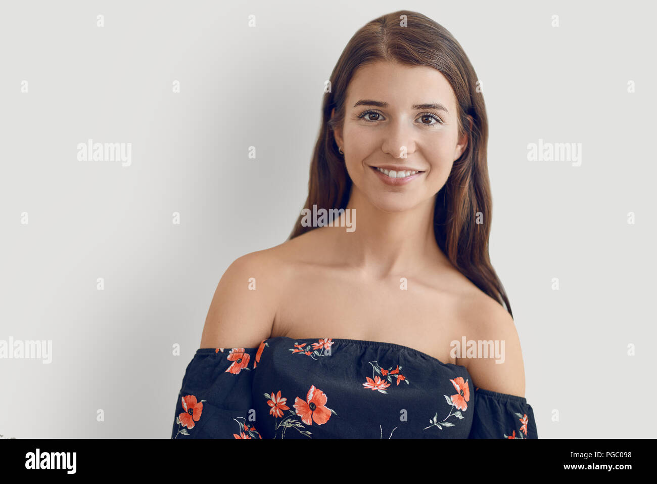 Attractive stylish woman in an off the shoulder summer dress with floral pattern standing against a white background with folded arms looking at the c Stock Photo