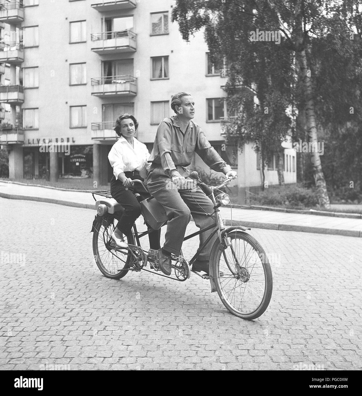 1950s couple on a tandem bicycle. Actor Kenne Fant and his wife Jeanine runs the neighbourhood cinema theatre and is riding their bike home after work. Notice the popular extra equipment mounted on the back. A small motor which gives an extra help forward. Sweden 1954  Photo Kristoffersson BD66-9 Stock Photo