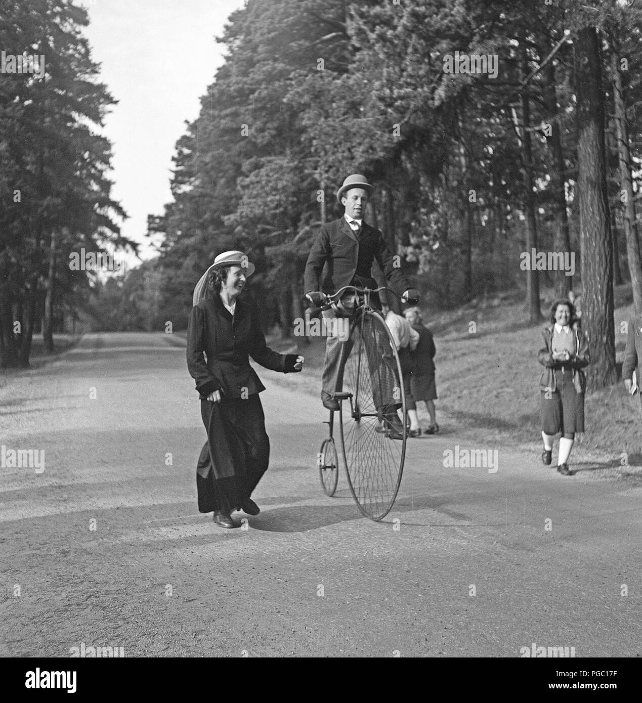 Penny farthing bicycle. A man is riding a penny-farthing bicycle and a woman is running by his side as if she is afraid he will fall. Sweden 1944 Photo Kristoffersson K131-2 Stock Photo