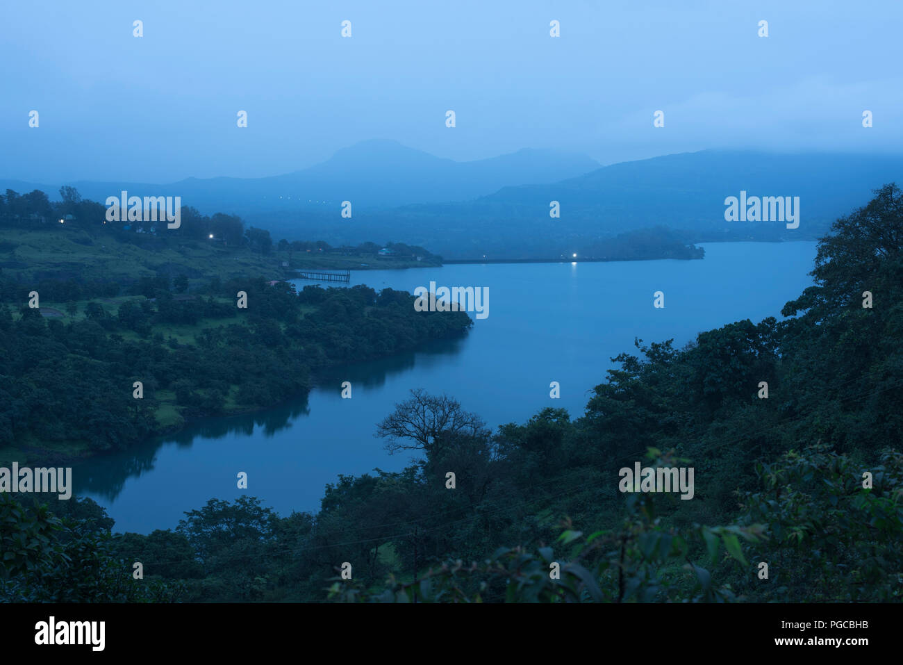Serene landscape at dawn with a lake at Bhandardara also known as Wilson Dam with dense forest and mountains in foggy monsoon atmosphere and lights Stock Photo