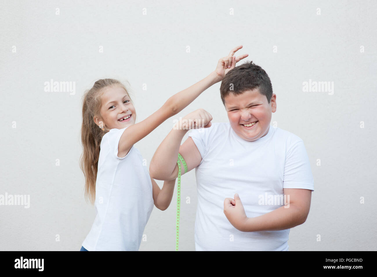 funny caucasian thin small girl measuring smiling fat boy muscle by tape fooling around Stock Photo