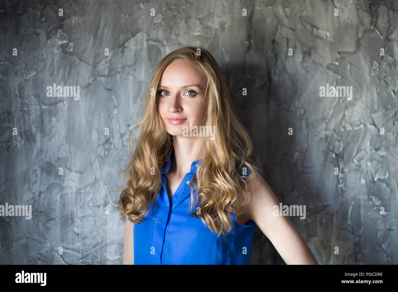 Portrait of the blonde woman in blue blouse staying in front of dark wall and keep smiling Stock Photo