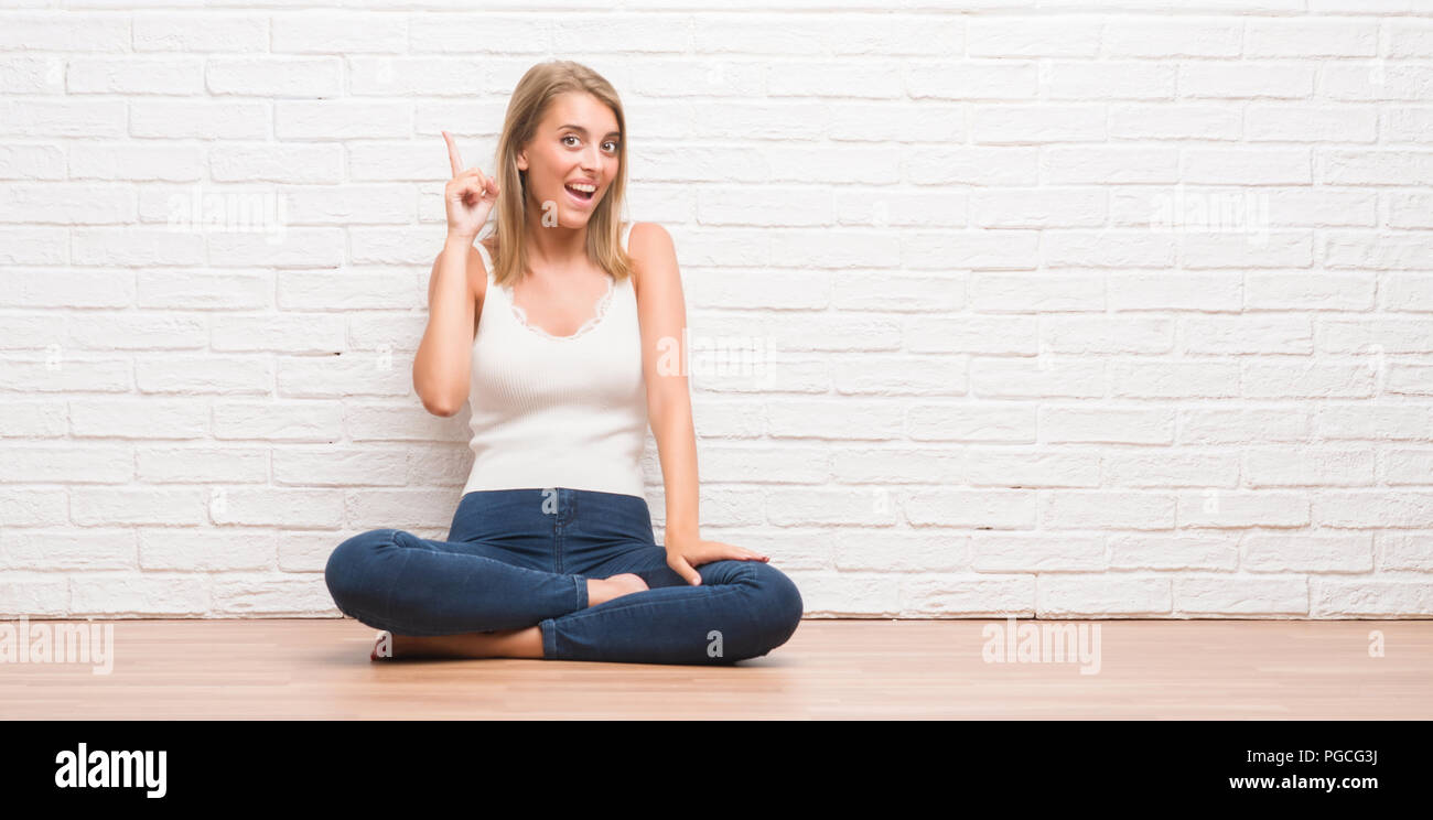 Beautiful young woman sitting on the floor at home pointing finger up ...