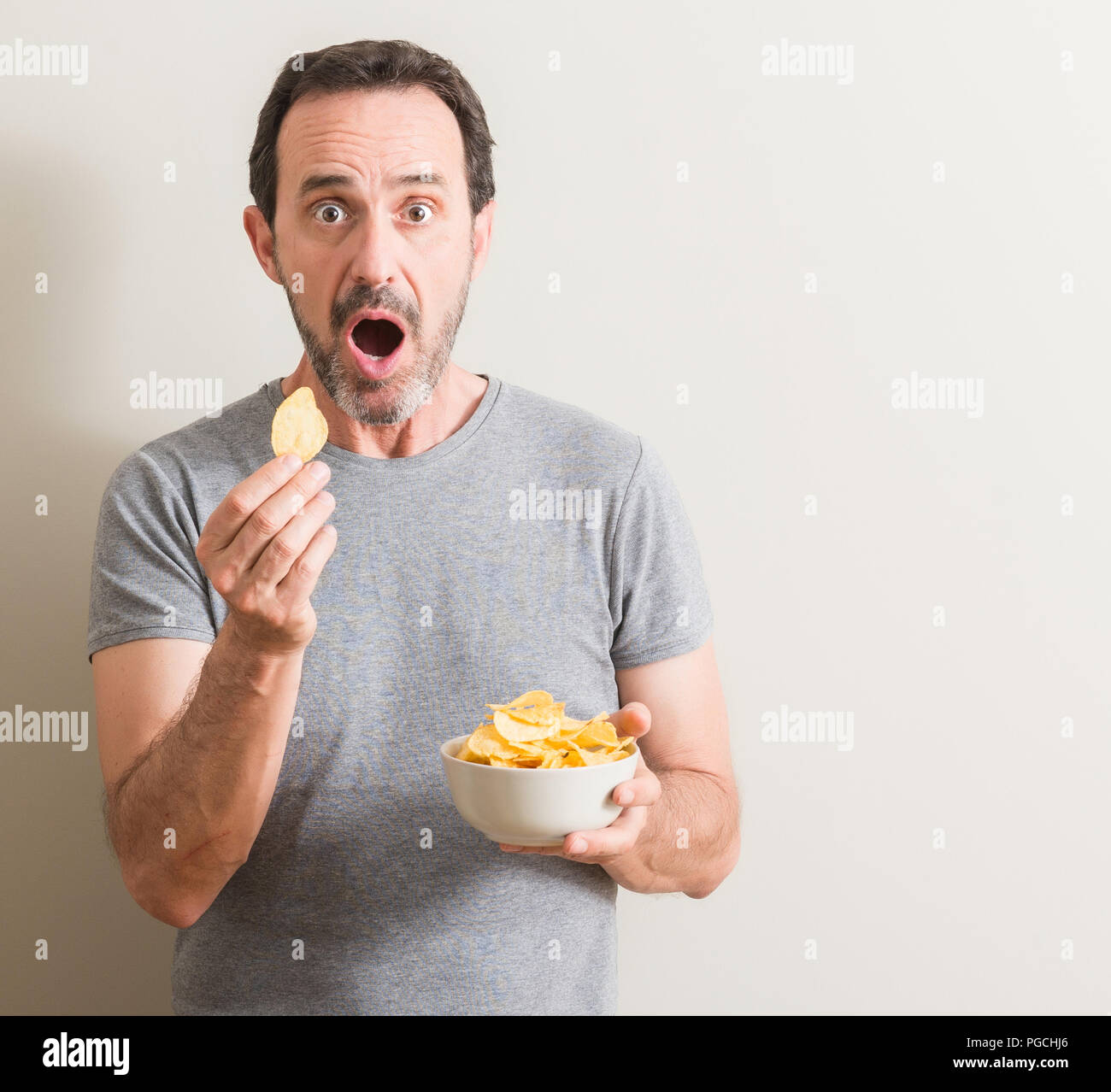 Senior man eating potato chips scared in shock with a surprise face