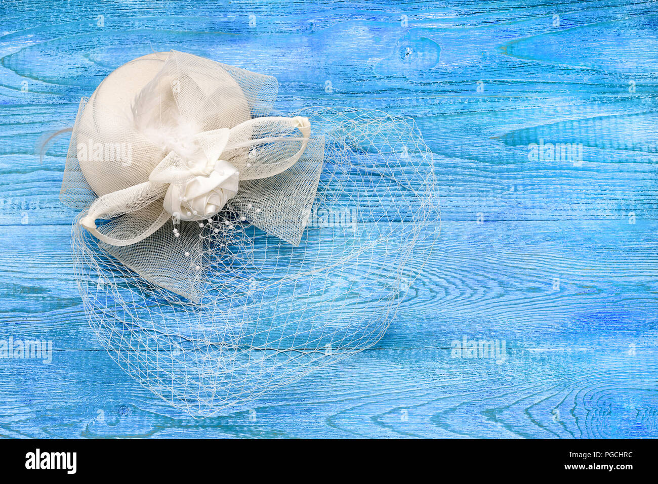A small white hat with a veil on a blue wooden table, a top view and a copy space. Decoration for the bride. Stock Photo