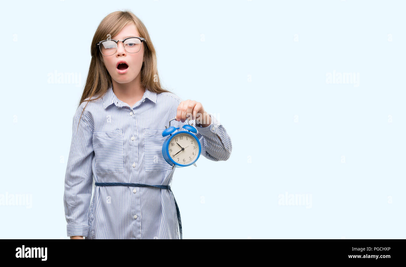 Young blonde child holding alarm clock scared in shock with a surprise