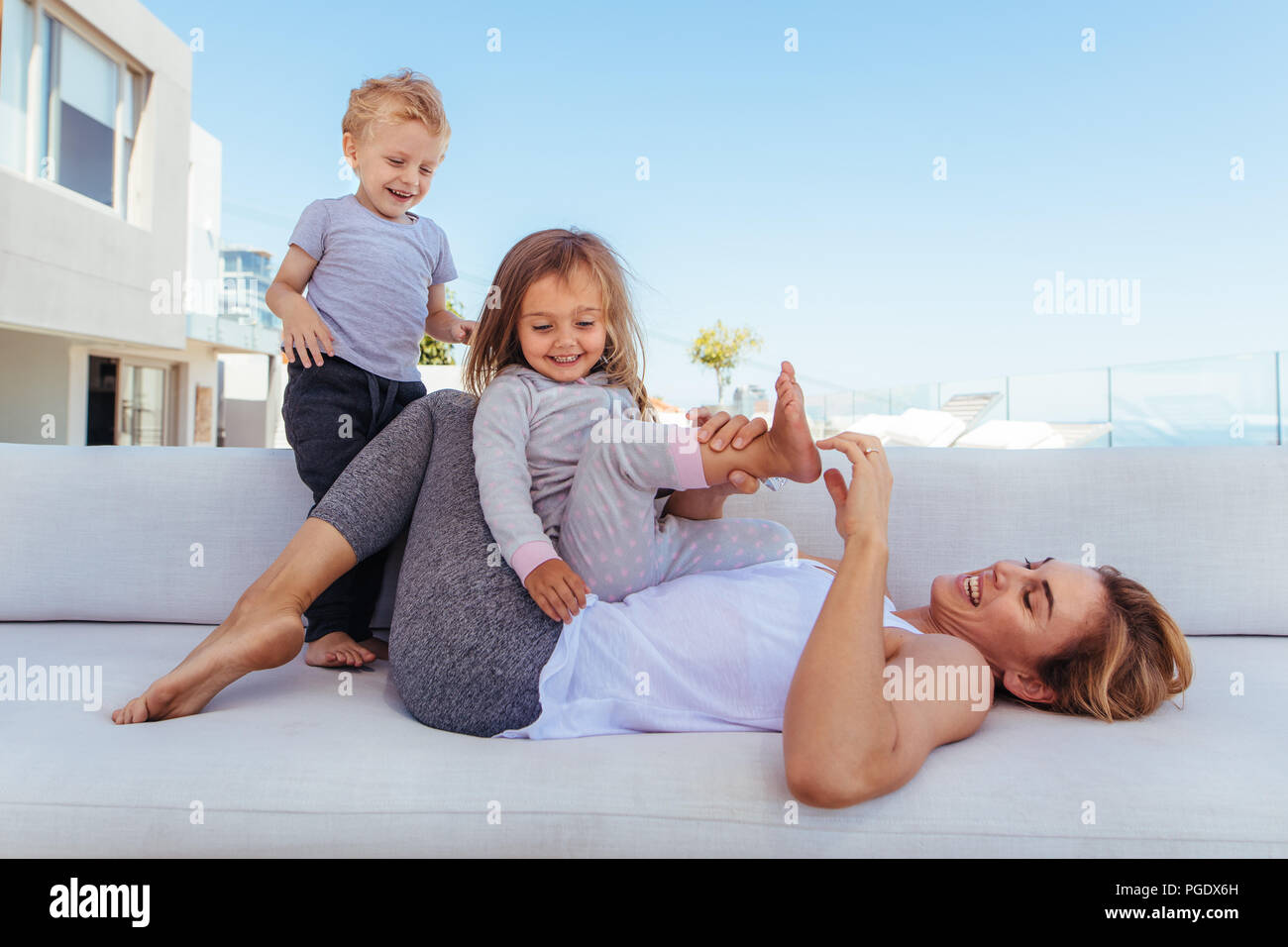 Young woman playing with her children on sofa at home. Happy family playing and spending time together at home. Stock Photo