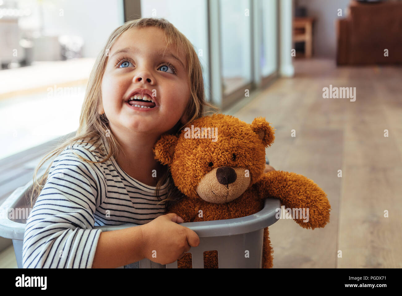 Adorable young girl sitting in a washing basket with her teddy bear. Beautiful girl child playing in a washing basket at home. Stock Photo