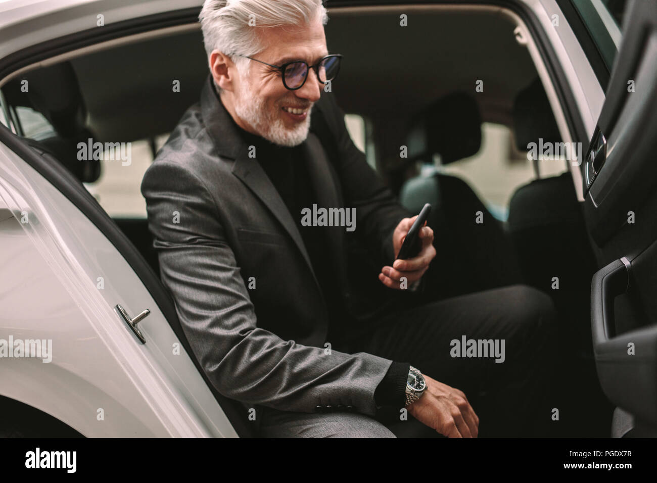 Smiling mature businessman sitting in the backseat of a taxi and looking at his phone. Senior male commuter taking a taxi ride in city. Stock Photo