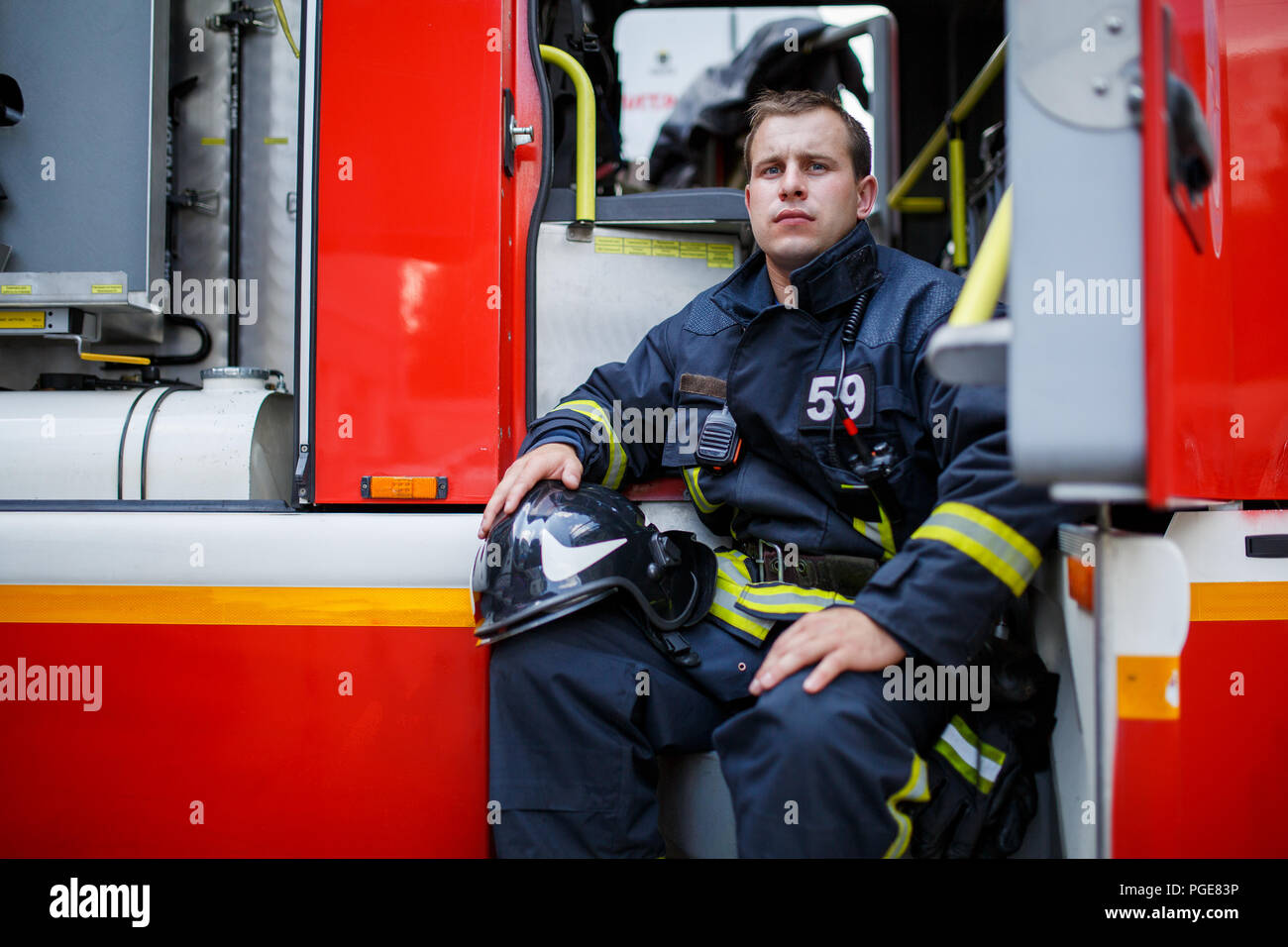 Photo of young fireman with helmet in overalls sits in fire truck Stock