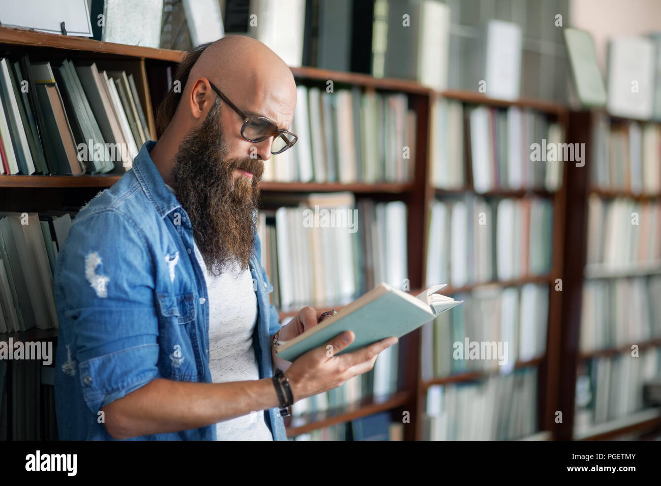 Handsome bearded man wearing glasses reading book in library Stock Photo