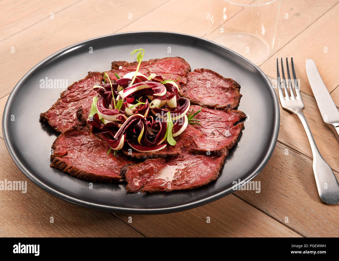 Sliced prime lean roast beef with radicchio salad topping on a black plate with utensils on a wooden table Stock Photo