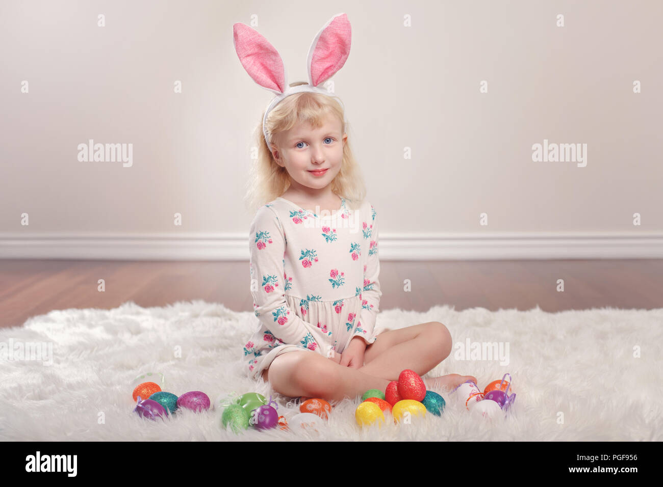 Cute adorable Caucasian child girl wearing Easter bunny rabbit ears sitting on soft fluffy rug carpet in studio. Kid holding holiday colorful eggs cel Stock Photo