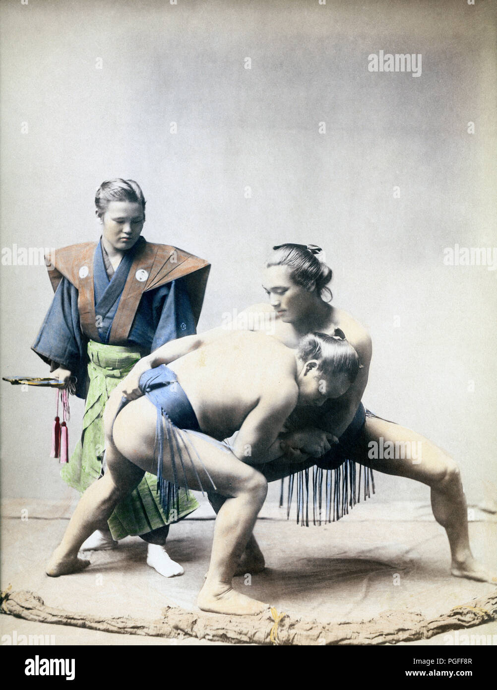 [ c. 1890s Japan - Sumo Wrestlers and Umpire ] —   Studio photograph of two rikishi (sumo wrestlers). In reality, the umpire would be wearing an eboshi (traditional black hat).  Albumen photograph published in 'Japan, Described and Illustrated by the Japanese', Shogun Edition edited by Captain F Brinkley. Published in 1897 by J B Millet Company, Boston Massachusetts, USA.  19th century vintage albumen photograph. Stock Photo