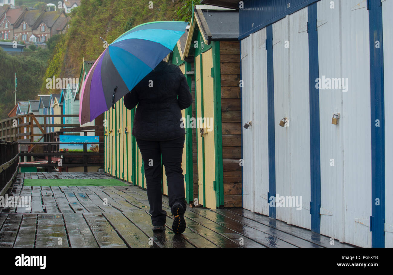 Beer, East Devon, UK. 26th August 2018.  UK Weather: The pretty beach at the fishing village of Beer was empty of holidaymakers as torrential downpours hit the south coast holiday resort over the bank holiday weekend. A woman with an umbrella walks near beach huts in the rain. Credit: DWR/Alamy Live News Stock Photo