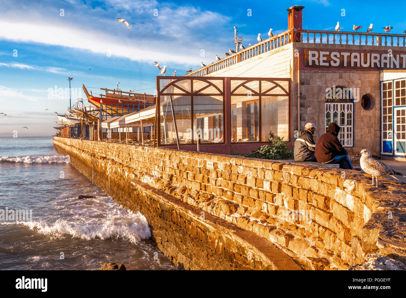 Morocco waterfront restaurant in Essaouira, a picturesque fishing village on the Atlantic coast. Waves hit a brick seawall. Beautiful light. Stock Photo