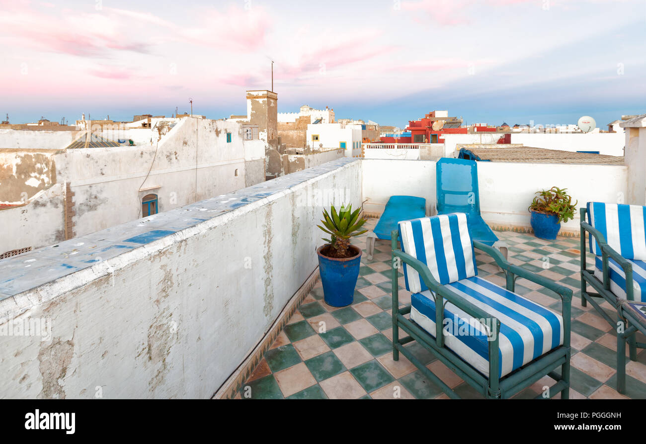 Rooftops in Essaouira, Morocco. Whitewashed buildings and pastel pink sky. Rooftop deck chairs with blue and white striped cushions in the foreground. Stock Photo