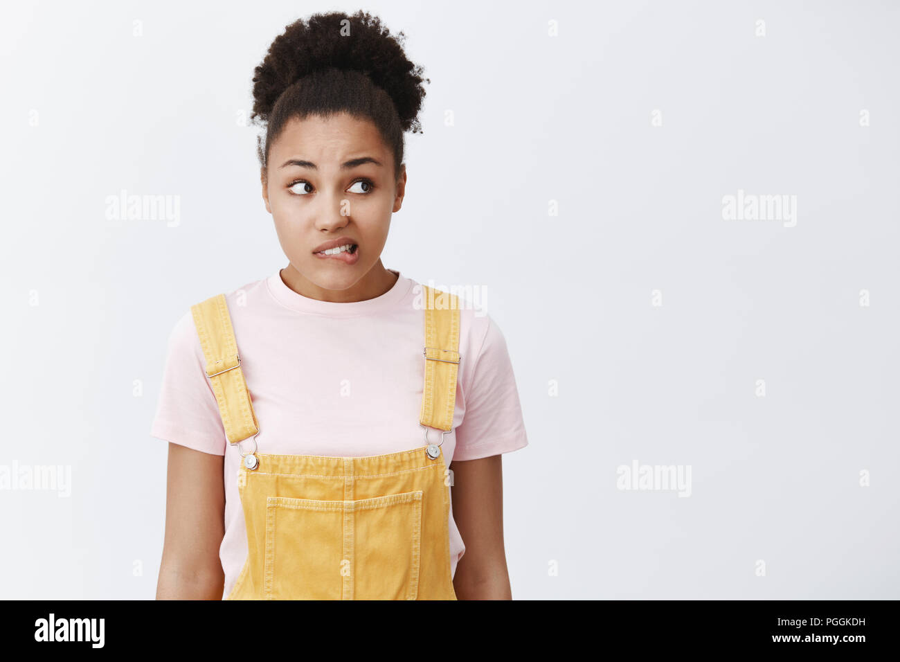 Woman feeling guilty, wanting say sorry. Portrait of nervous and worried cute African American girl in yellow overalls, biting lip and gazing anxiously right, standing over gray background Stock Photo