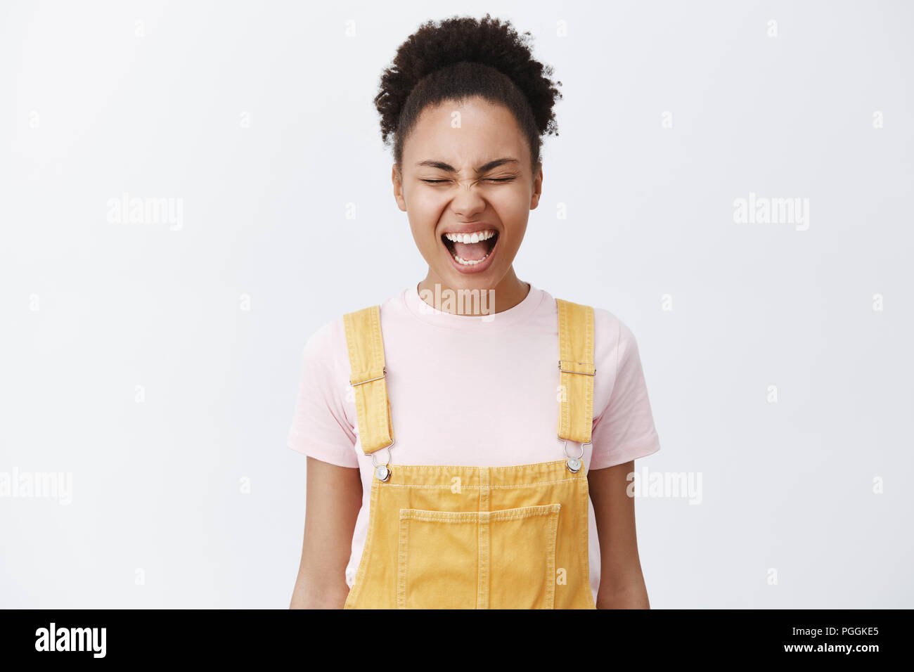 Letting emotions out feels good. Careless beautiful African American with smooth and clean skin in yellow overalls, shouting with closed eyes, releasing stress, trying calm down over grey wall Stock Photo