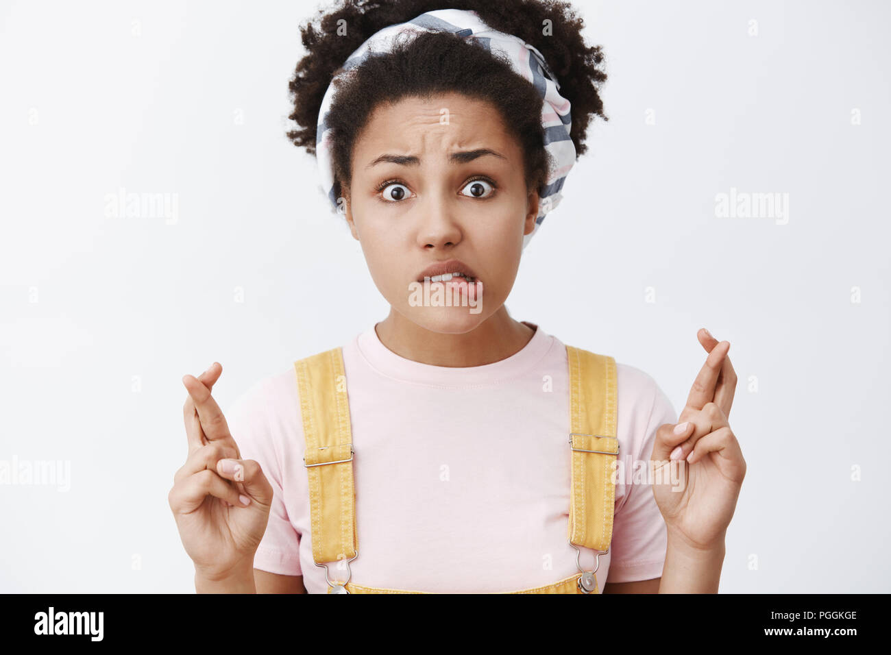 Girl prayes and hopes boyfriend will call after date. Portrait of worried intense charming dark-skinned female in dungarees and headband, biting lip and crossing fingers in hope gesture Stock Photo