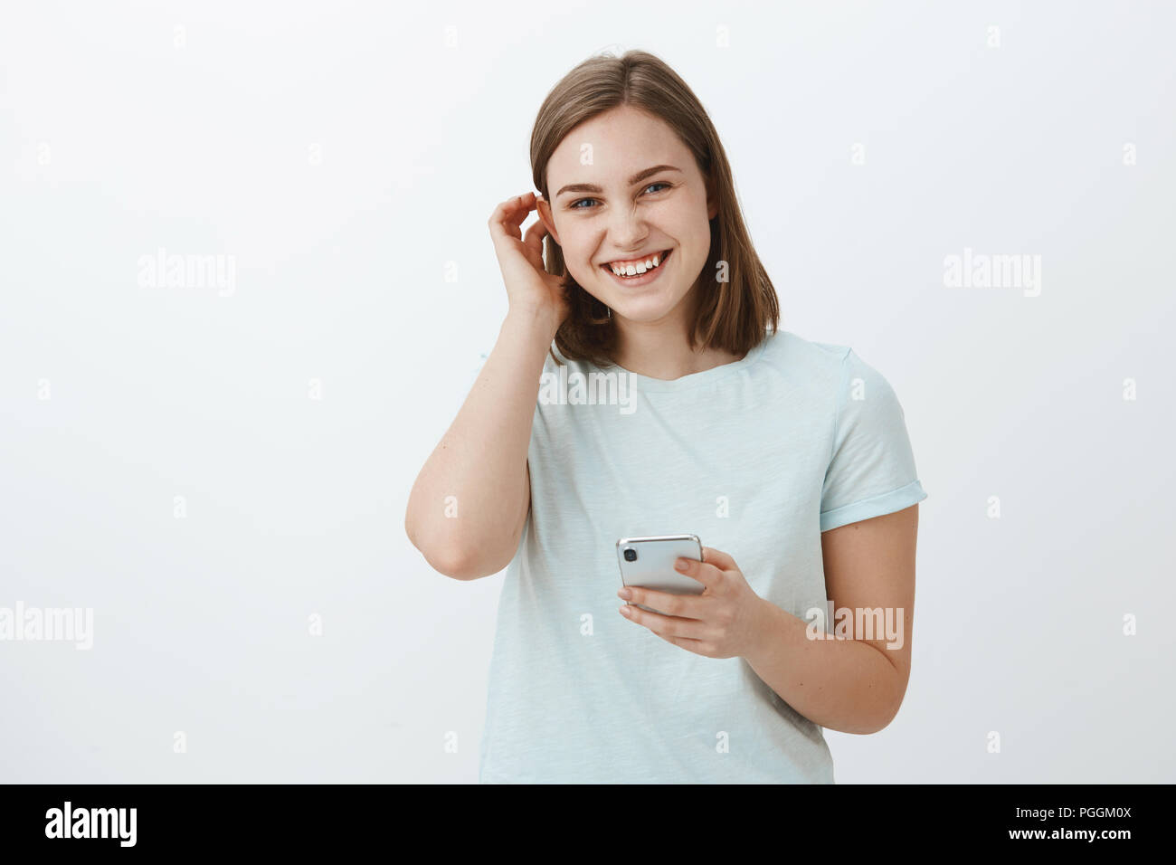 Woman exchanging numbers with cute guy on festival. Charming friendly-looking young girl flicking hair behind ear flirty and laughing while gazing at camera holding smartphone against gray wall Stock Photo